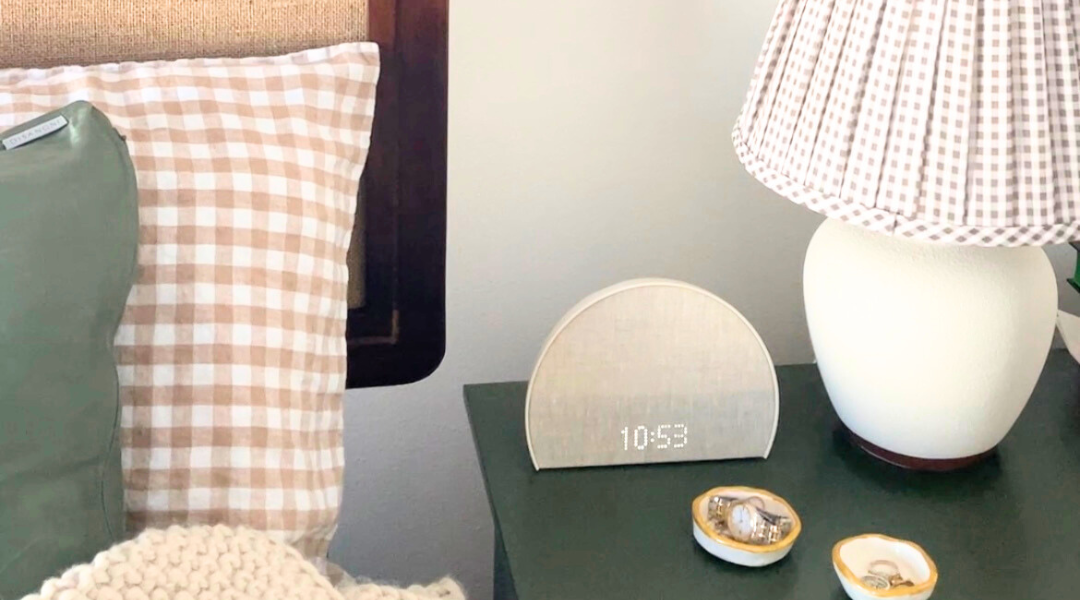 A made bed in the sunlight with an alarm next to it on the table with a lamp