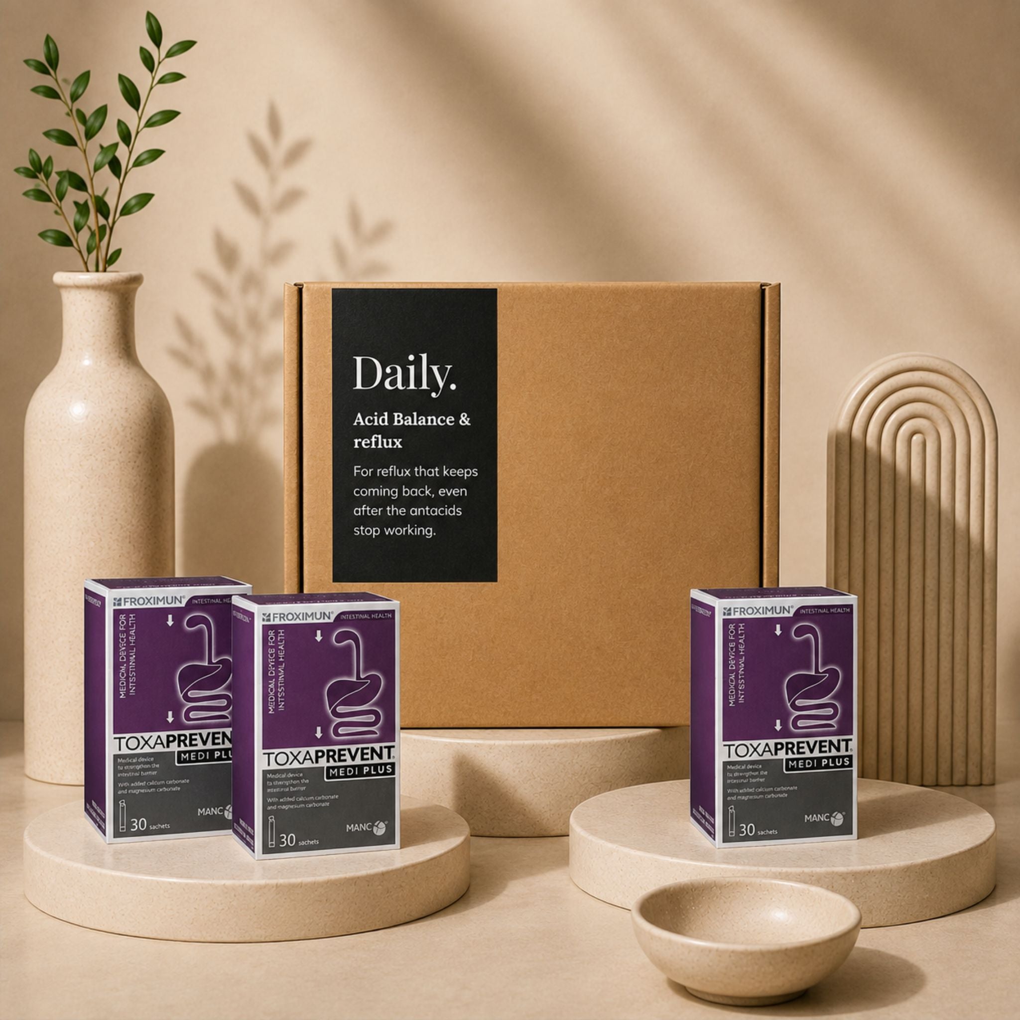 A styled product display featuring a large brown cardboard box labeled 'Daily. Acid Balance & reflux' behind three purple and white 'Toxaprevent Daily' supplement boxes, arranged on beige pedestals with a ceramic vase, plant, and sculptural decor in a warm neutral setting.