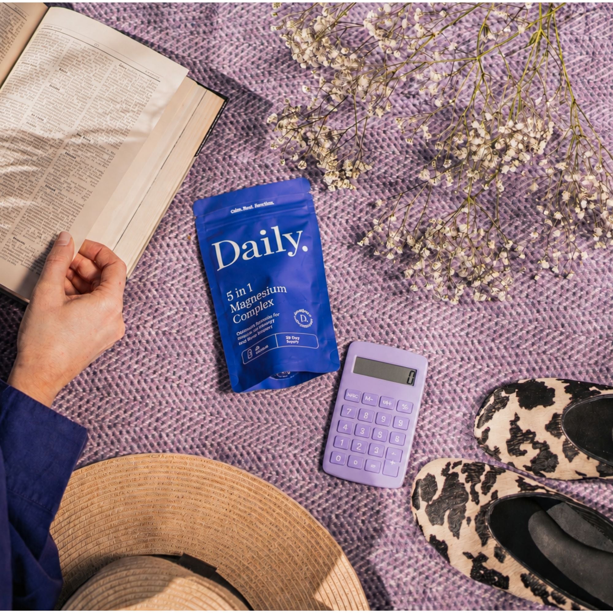A blue Daily 5 in 1 Magnesium Complex supplement pouch is displayed in a styled flat-lay scene on a textured purple blanket, surrounded by an open book, dried flowers, a lavender calculator, leopard-print flats, and a straw hat.