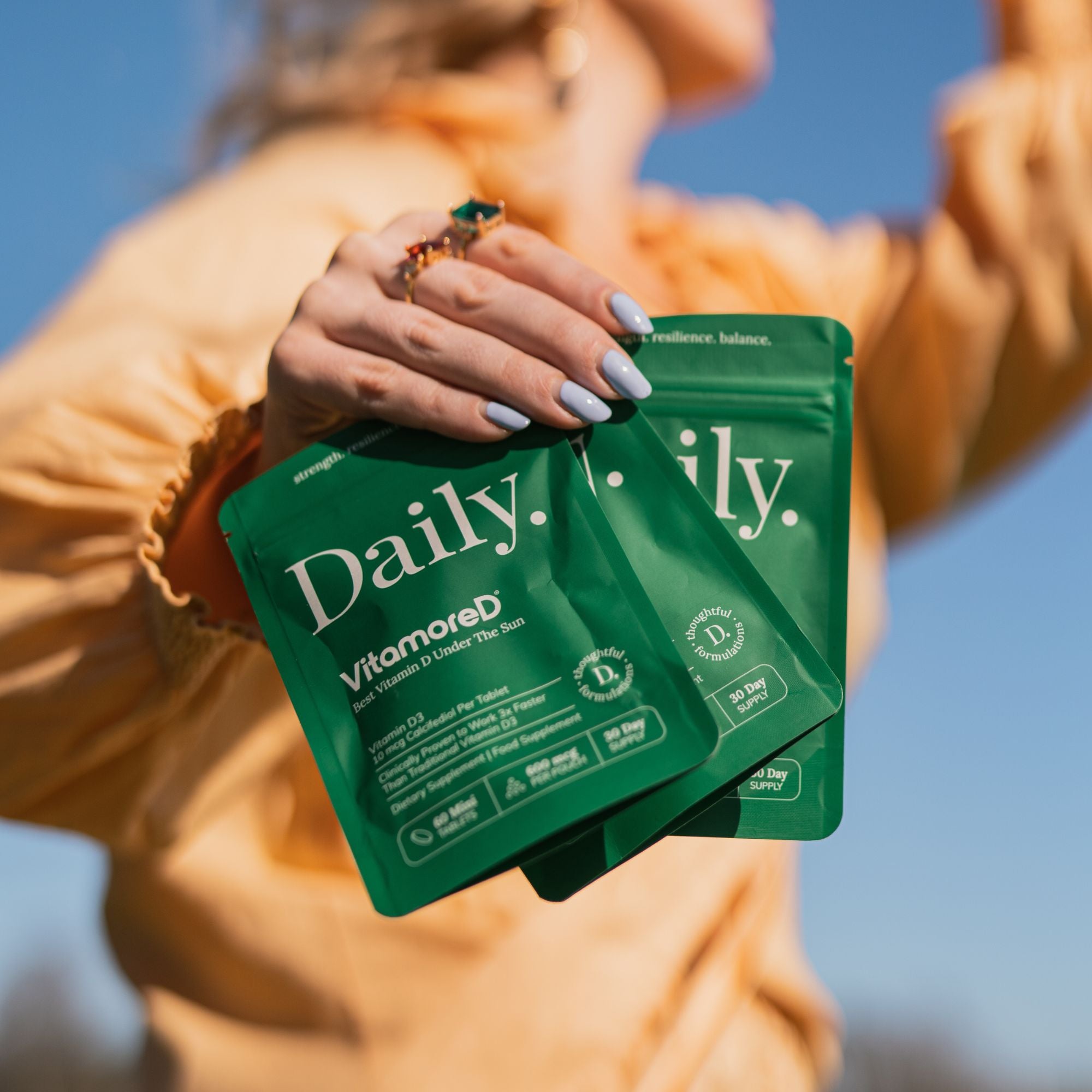 A person outdoors holds three green Daily Vitamored skincare pouches in front of their face, with a bright blue sky in the background.