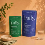 Two Daily brand supplement pouches standing on a warm beige surface: a green pouch labeled VitamoreD on the left and a blue pouch labeled 5 in 1 Magnesium Complex on the right. A small bowl of capsules, a glass of water, and a plant branch are arranged around them in a styled studio setup.