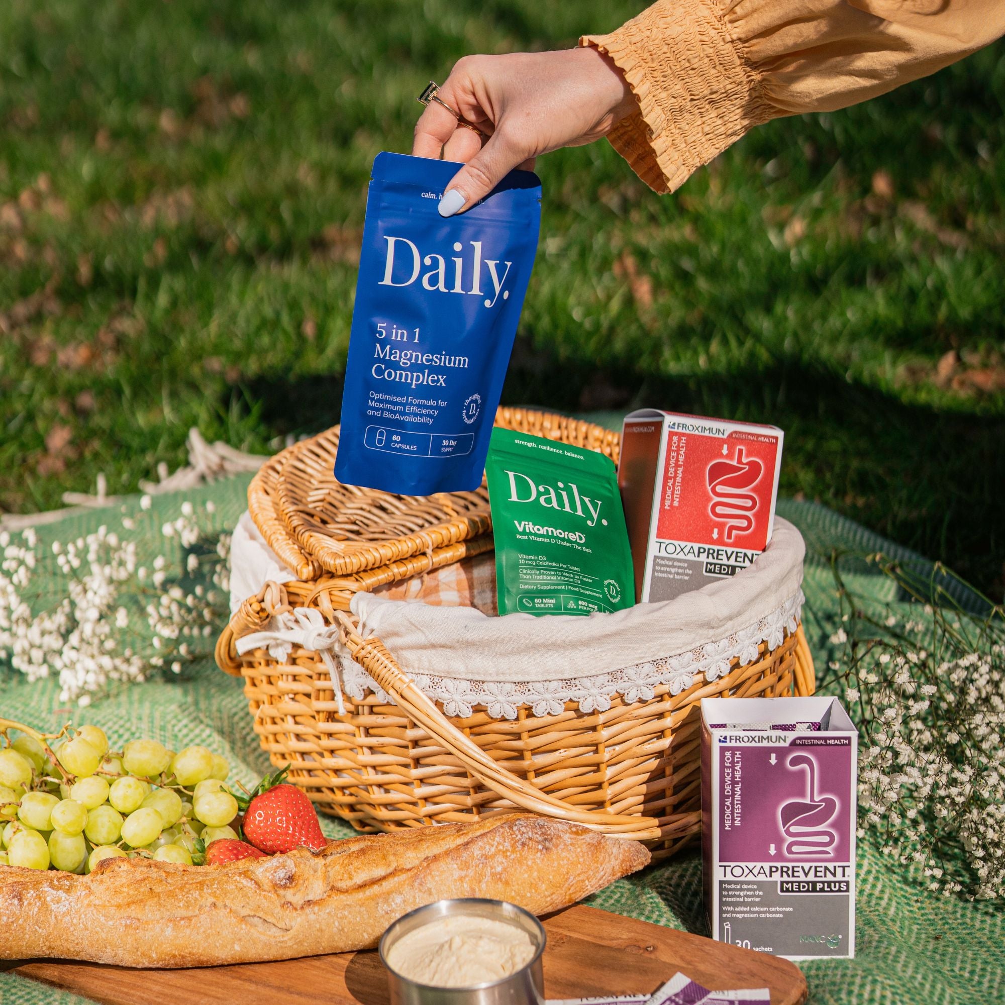 A hand holds a blue Daily 3 in 1 Magnesium Complex packet above a wicker picnic basket on a green blanket outdoors. The basket contains other Daily branded packets, including Vitamin and Toxapreven, with a baguette, grapes, strawberry, and bread on a wooden board in the foreground.