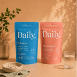 Two Daily. branded stand-up pouches, one blue for Dolvent and one coral pink for Electrolytes, displayed on a warm beige surface with a glass of water, a bowl of tablets, and a plant in the background.