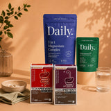 A styled product display of three 'Daily.' supplement packages and two boxed supplements on a warm beige surface with a matching backdrop.