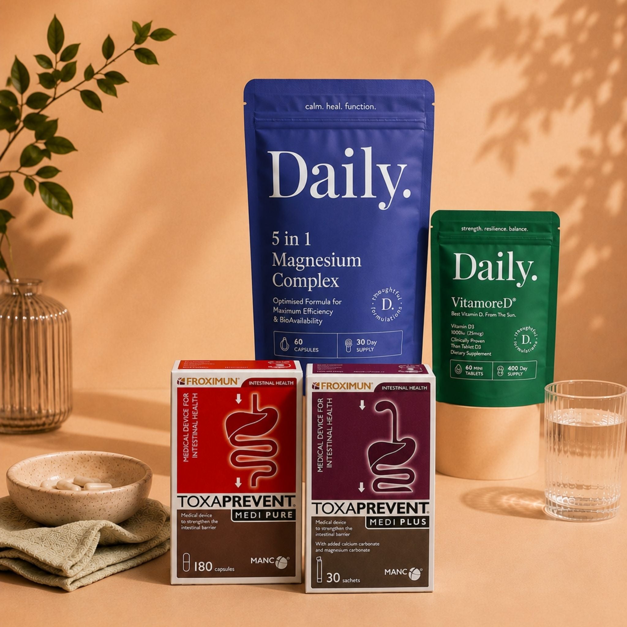 A styled product display of three 'Daily.' supplement packages and two boxed supplements on a warm beige surface with a matching backdrop.