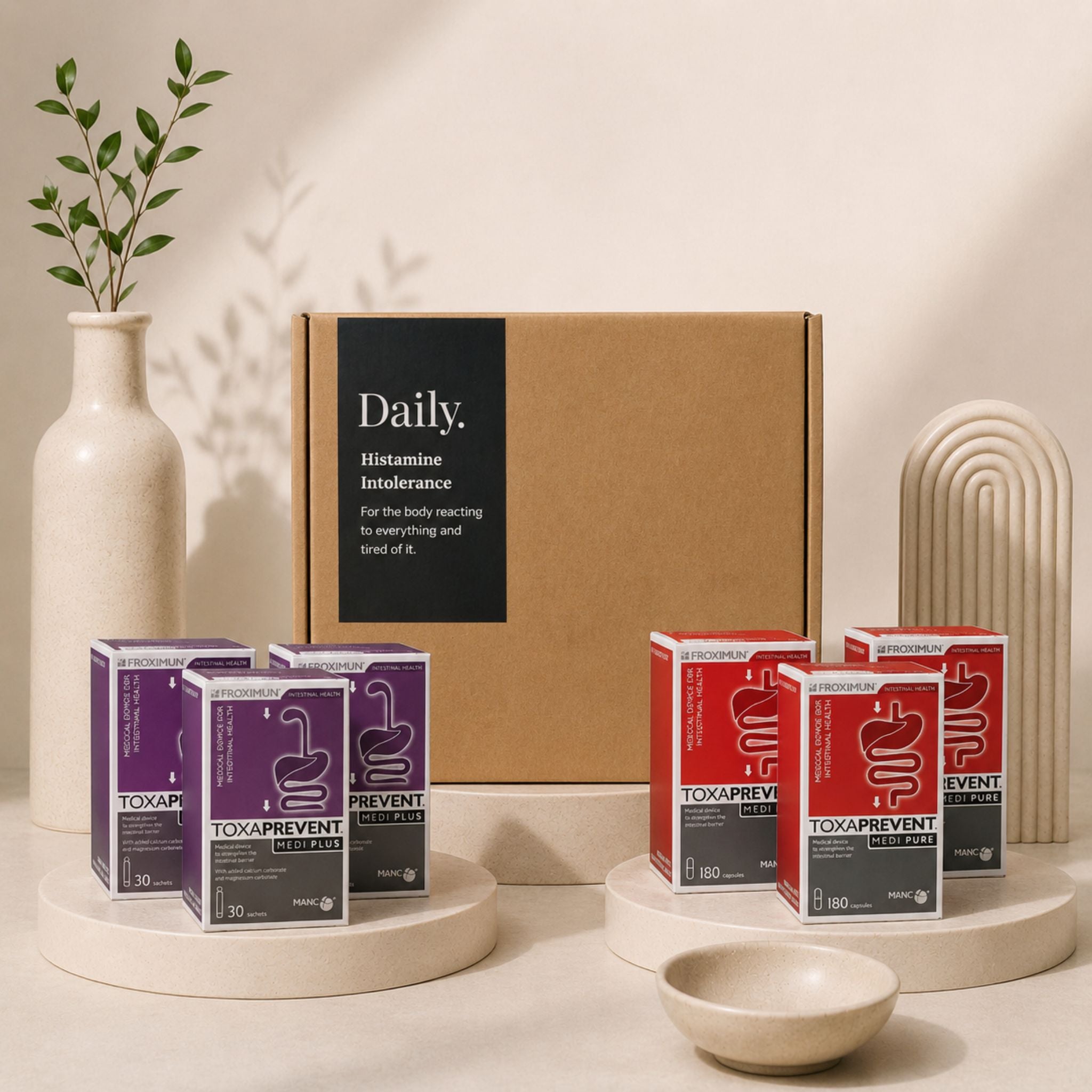 A styled product display featuring a large brown cardboard box labeled 'Daily Histamine Intolerance' behind several smaller product boxes in purple and red packaging, arranged on round pedestals with a ceramic vase, plant, and small bowl in a warm neutral setting.