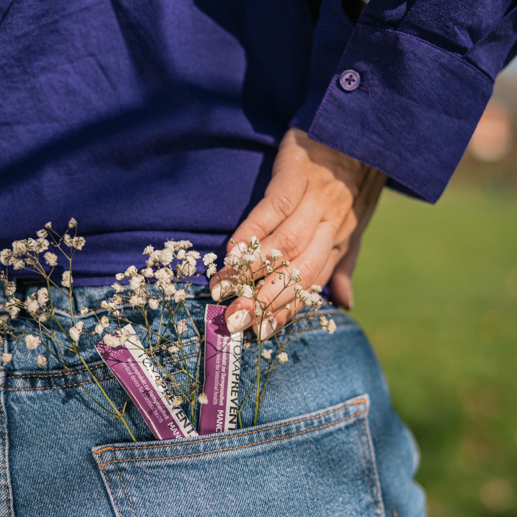 A close-up lifestyle shot of a person wearing blue jeans and a dark purple shirt, with baby's breath flowers tucked into the back pocket alongside two purple packets of Toxaprevent zeolite powder.